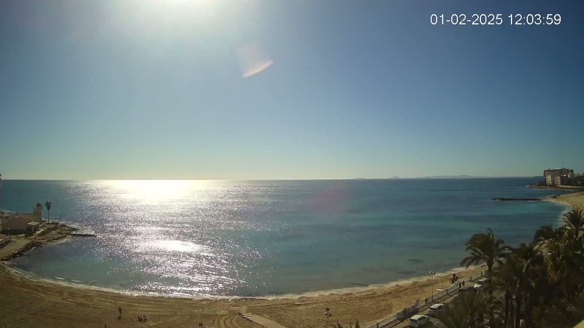 A sunny day at a beach with calm, sparkling water, a sandy shore, and palm trees along a roadway.