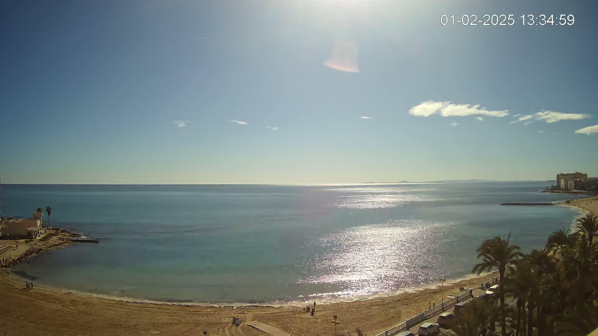 A sandy beach curves around a calm, sunlit turquoise sea under a mostly clear blue sky.