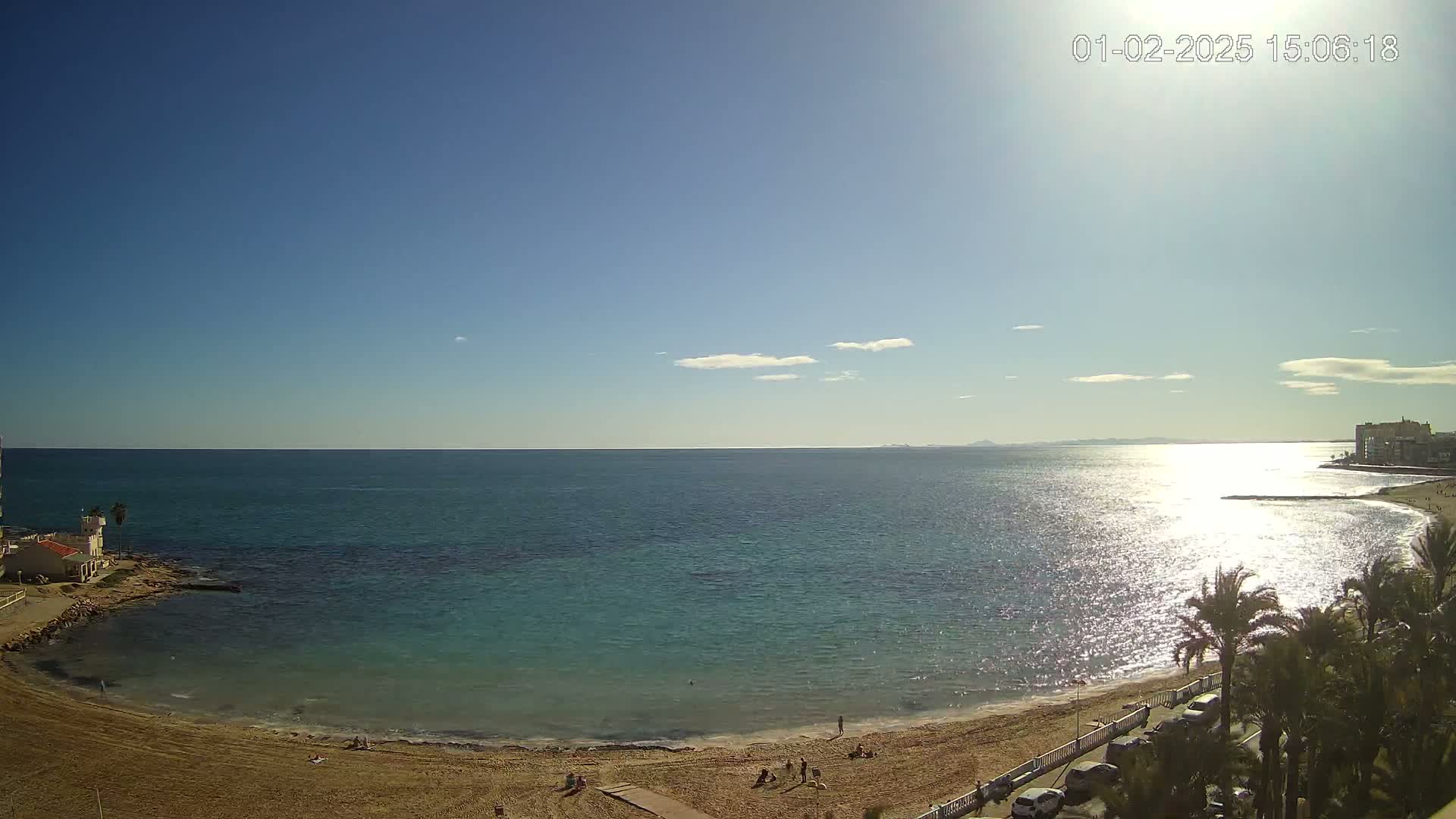 A sunny day at a beach shows a calm, teal sea with a sandy shore, palm trees, and buildings in the distance.