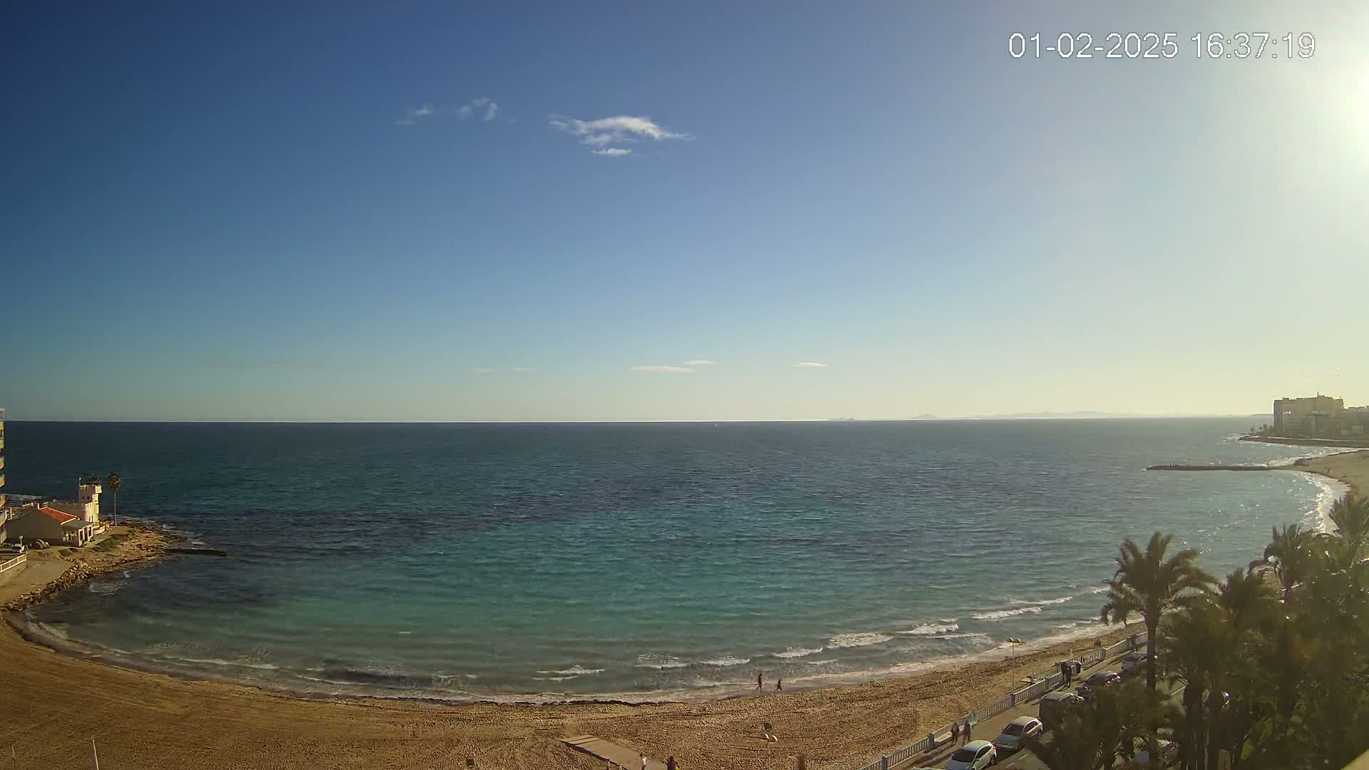 A sandy beach curves along a calm, teal ocean under a mostly clear blue sky.