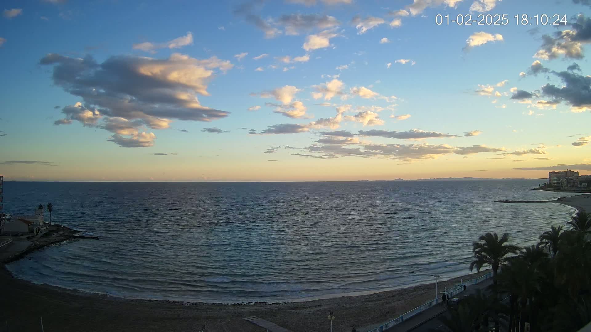 A calm sea with small waves laps a sandy beach under a partly cloudy sunset sky.