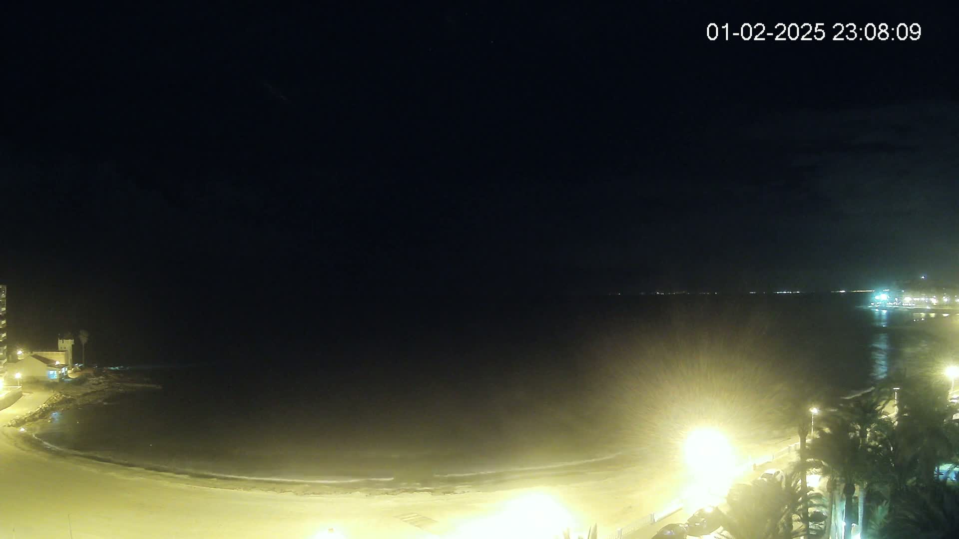 A nighttime view of a sandy beach and calm ocean under a clear, dark sky, illuminated by streetlights along the shore.