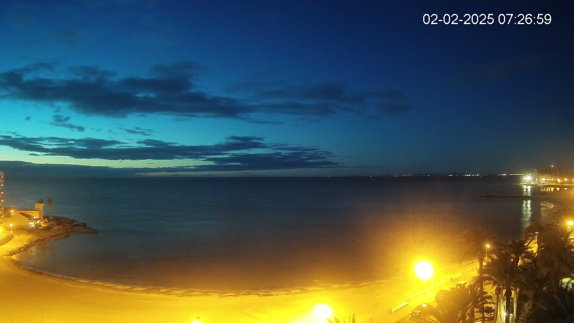 A nighttime beach scene shows a calm, dark-blue ocean under a mostly clear, twilight sky, with illuminated palm trees and a sandy shore.