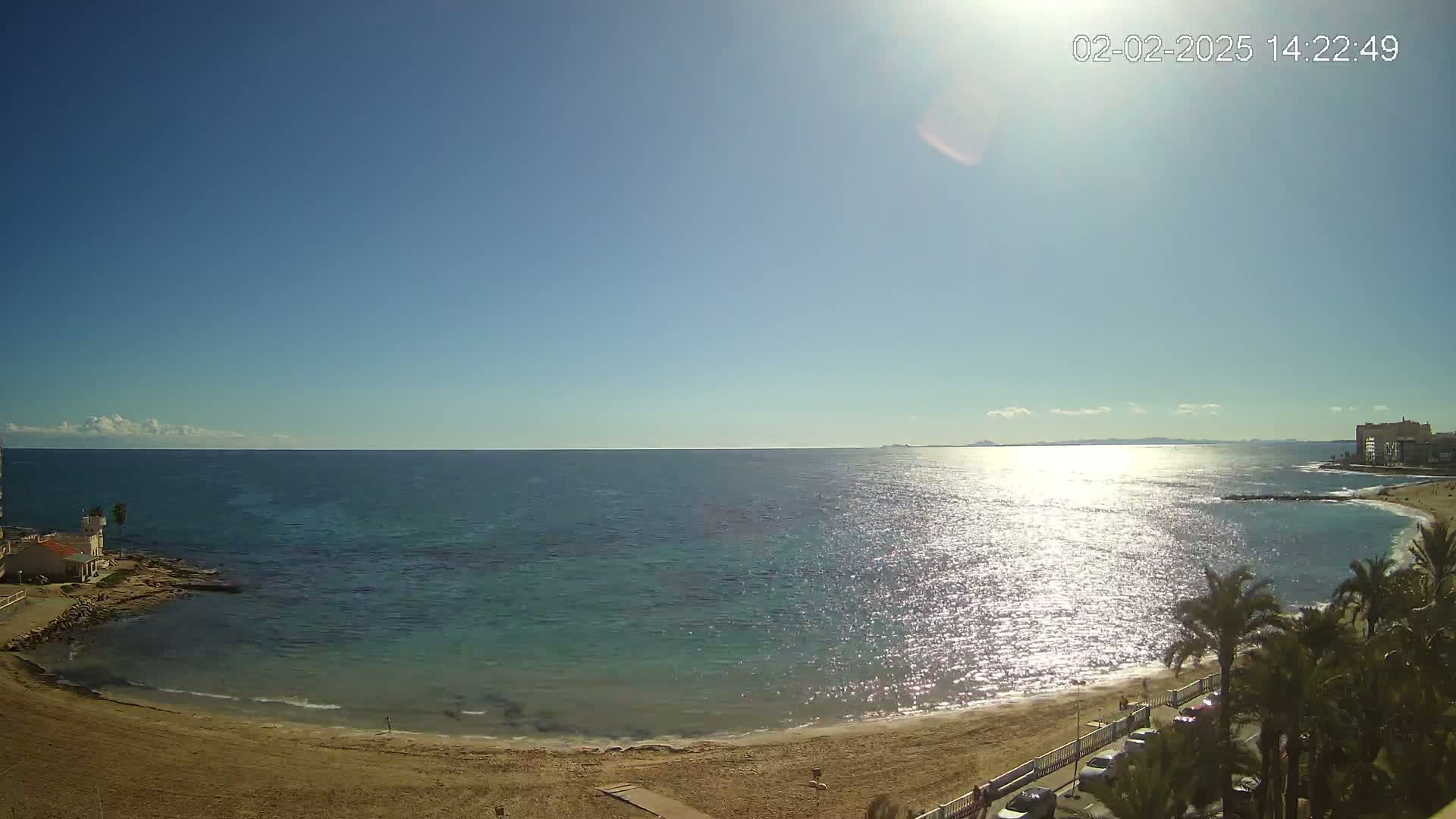 A sandy beach curves around a calm, sunlit sea under a clear blue sky.