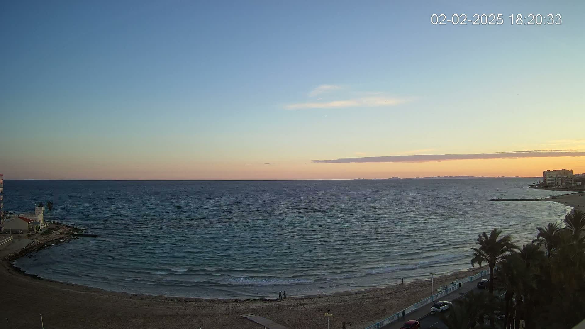 A calm sea at sunset with a sandy beach, palm trees, and buildings in the distance under a clear, pale pink and orange sky.