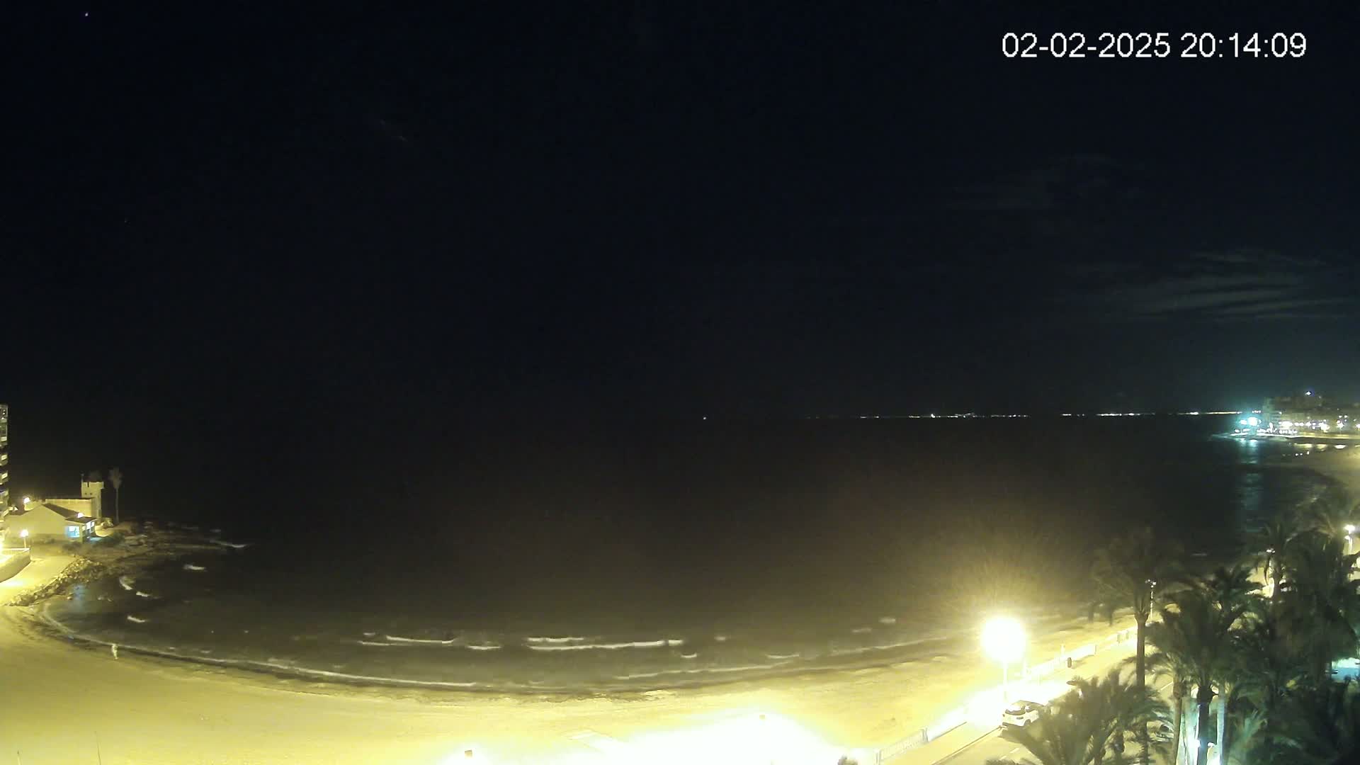 A nighttime view of a sandy beach with gentle waves under a dark, mostly clear sky, illuminated by streetlights and the distant lights of buildings.