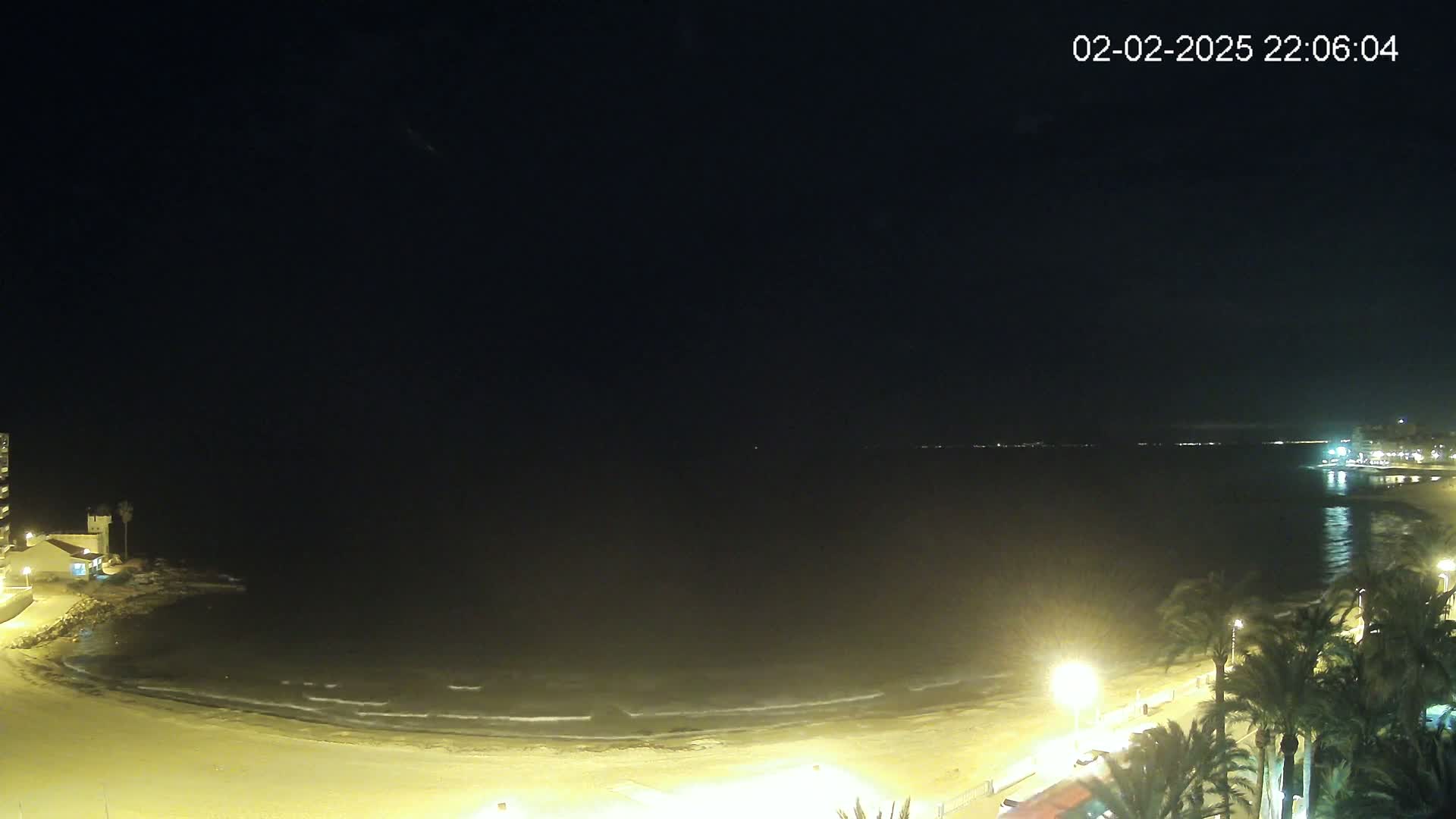 A nighttime beach scene shows gently lapping waves on a sandy shore, palm trees lining a paved walkway, and buildings in the distance under a dark sky.
