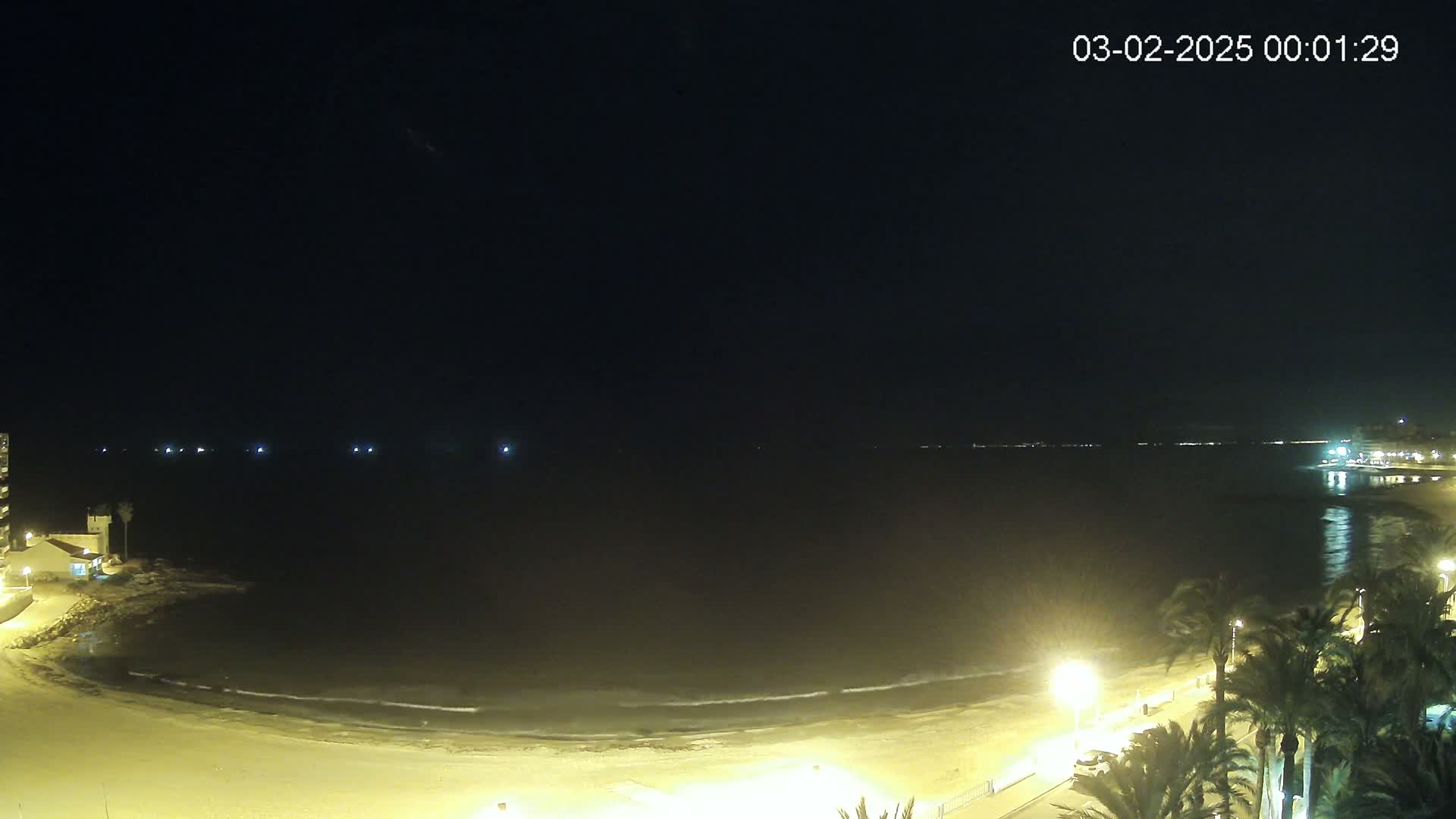 A nighttime beach scene shows gently lapping waves on a sandy shore, illuminated by streetlights and buildings, under a clear, dark sky.