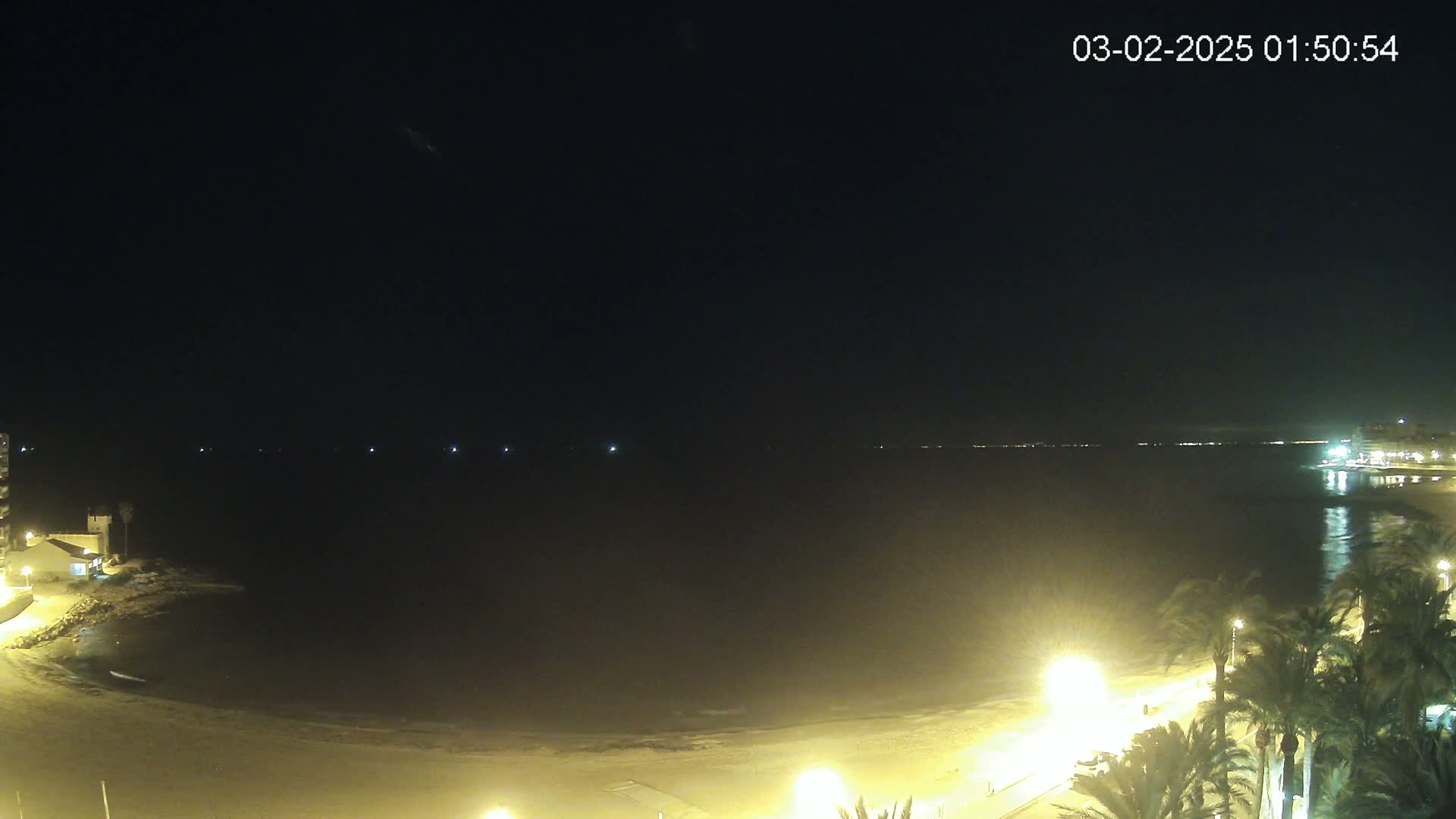 A nighttime view of a sandy beach and calm ocean, illuminated by streetlights and buildings along the shore.
