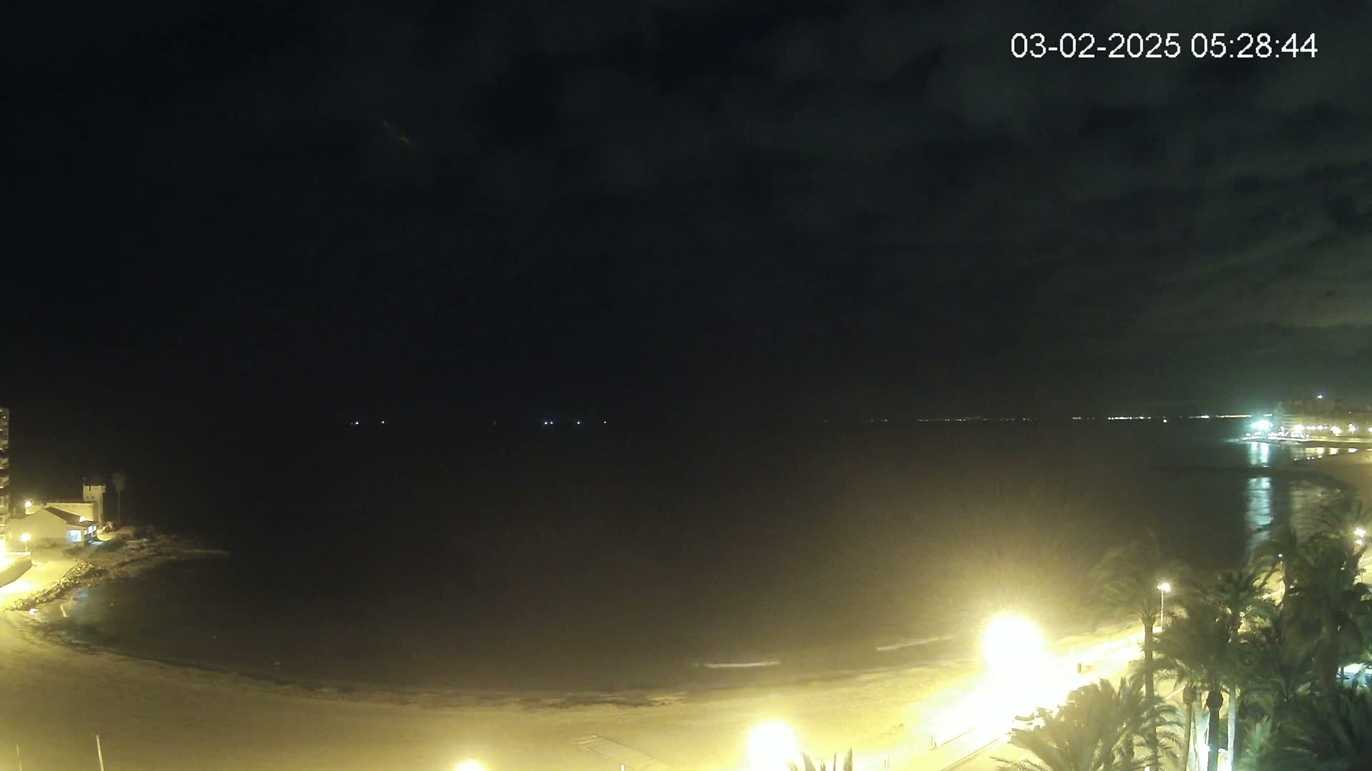 A nighttime view of a beach with calm water, illuminated by streetlights and buildings along the shoreline under a partly cloudy sky.