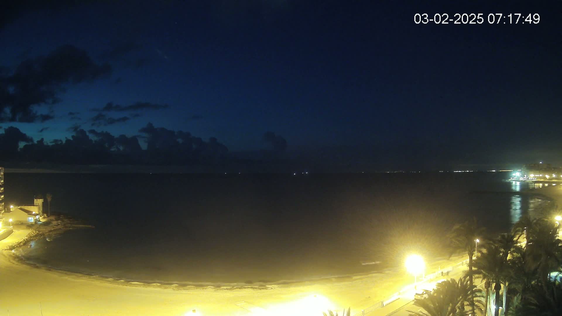 A night-time coastal scene shows a dark sea under a mostly clear, dark blue sky with some clouds, a sandy beach illuminated by artificial lights, and palm trees lining a walkway.
