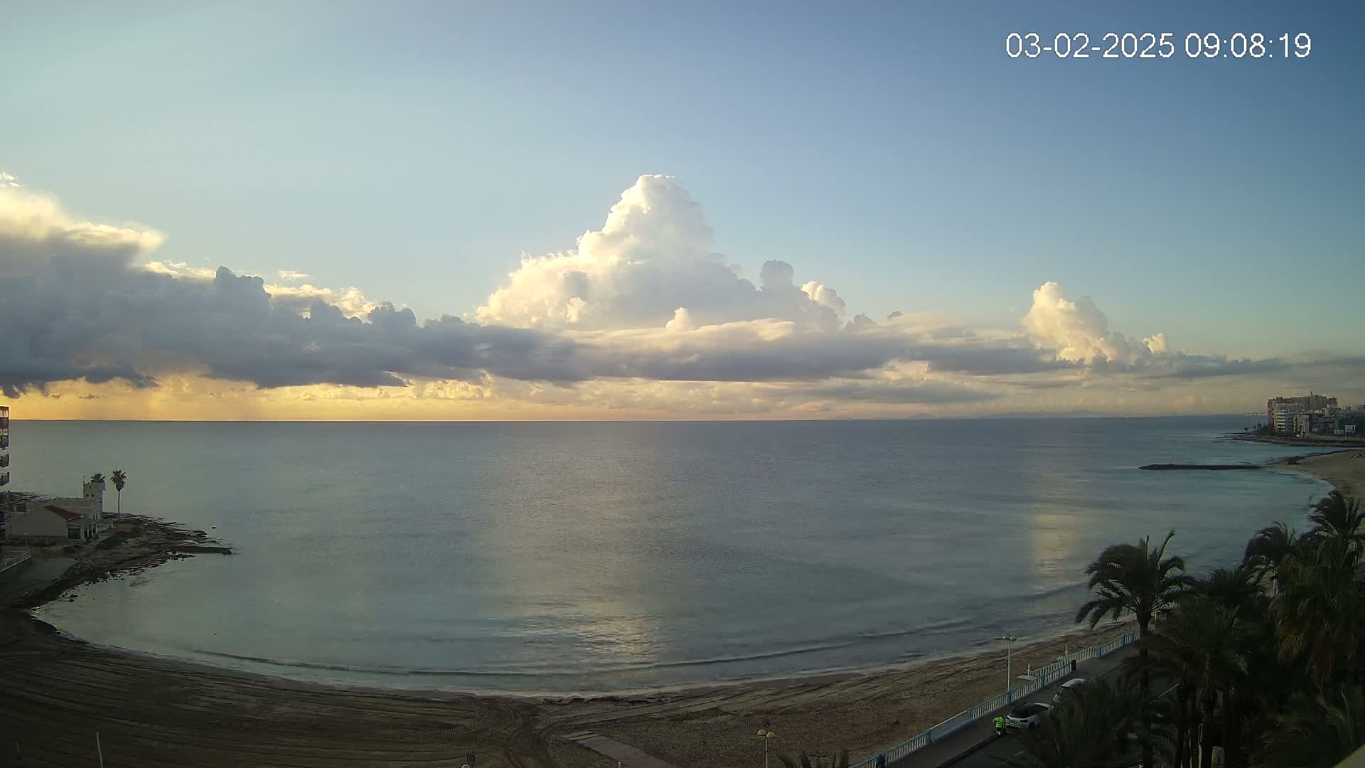 A calm ocean meets a sandy beach under a partly cloudy sky at sunrise.