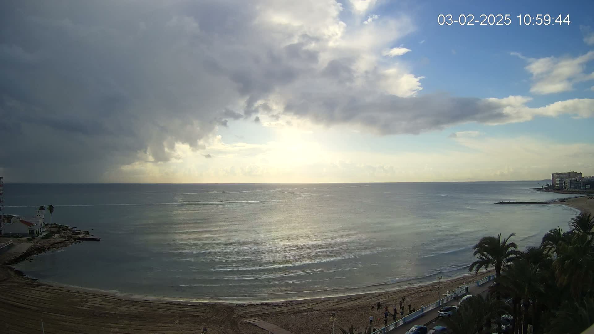 A partly cloudy day shows a mostly calm ocean meeting a sandy beach lined with palm trees and buildings in the distance.