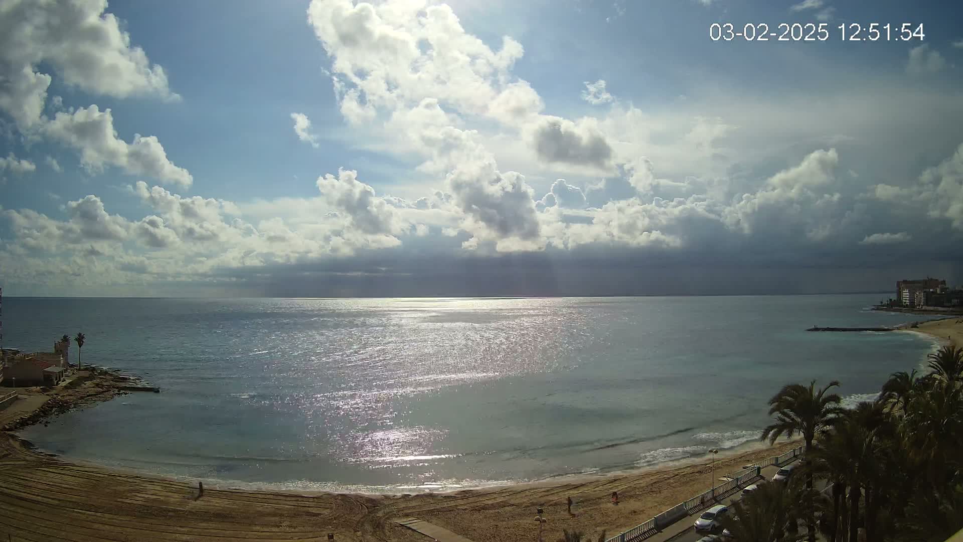 A partly sunny day shows a sandy beach and calm ocean with a few buildings visible in the distance, under a sky filled with fluffy cumulus clouds.