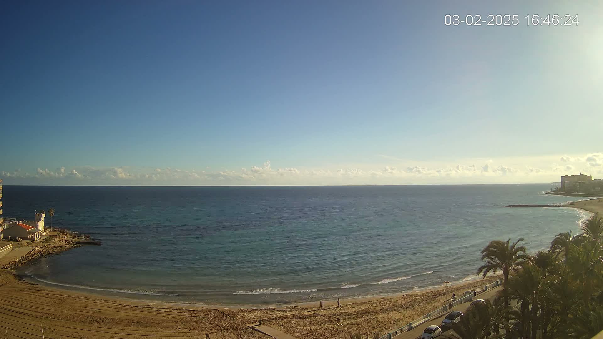 A sandy beach curves around a calm, turquoise sea under a mostly clear blue sky with a few wispy clouds.