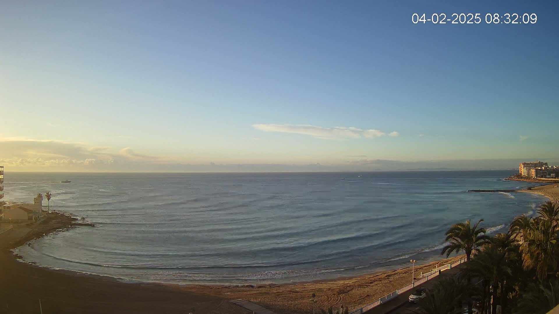 A sandy beach curves gently into a calm sea under a mostly clear sky, with a few wispy clouds and buildings visible in the distance.