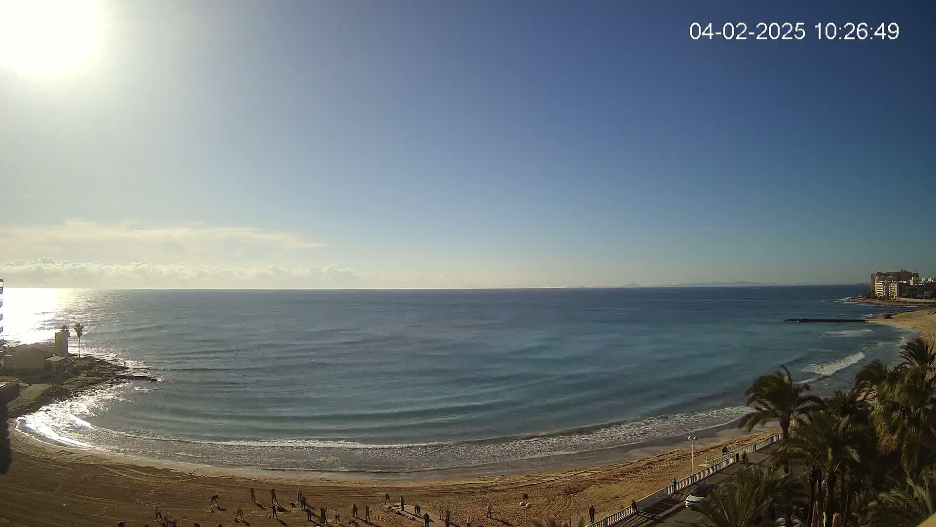 A sandy beach curves along a calm, clear ocean under a bright, sunny sky.