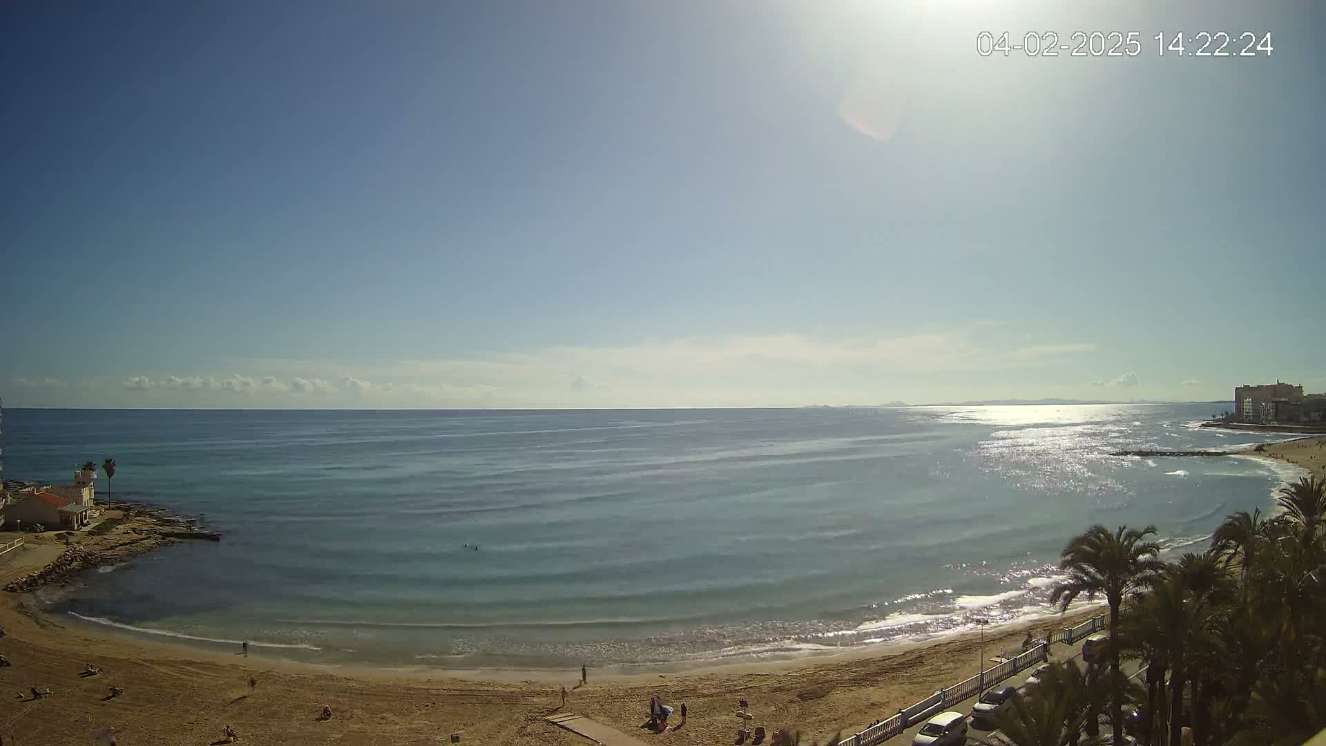 A sunny day at a sandy beach with calm, light-blue water, palm trees, and a few people are visible.