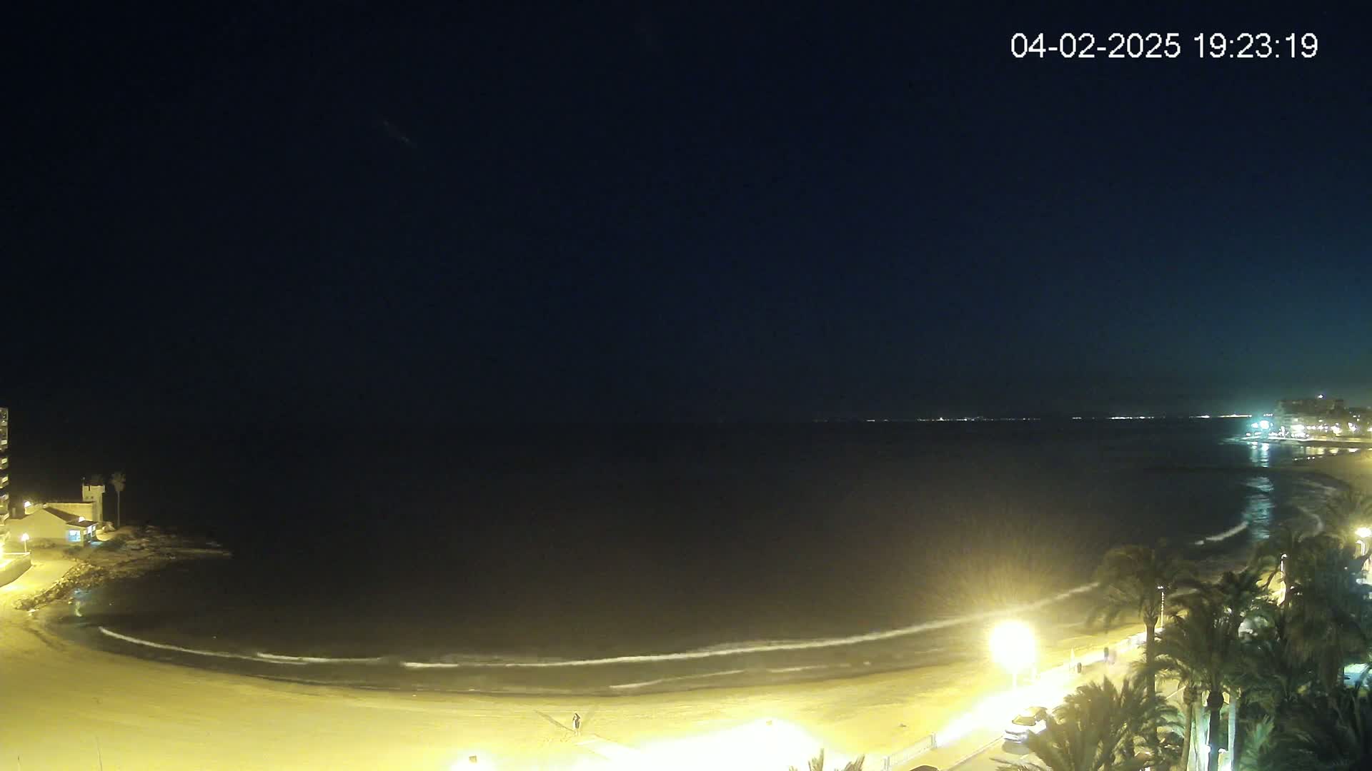 A nighttime view of a calm ocean beach with gentle waves lapping the shore, illuminated by streetlights and buildings along the coastline.