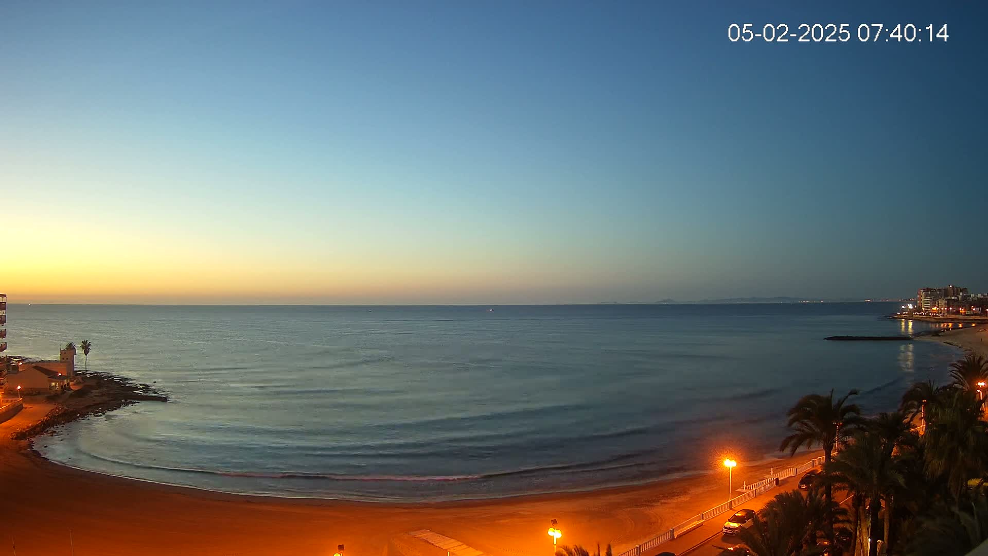 A sandy beach at dawn with calm ocean waves, palm trees lining a street, and a building visible in the distance.