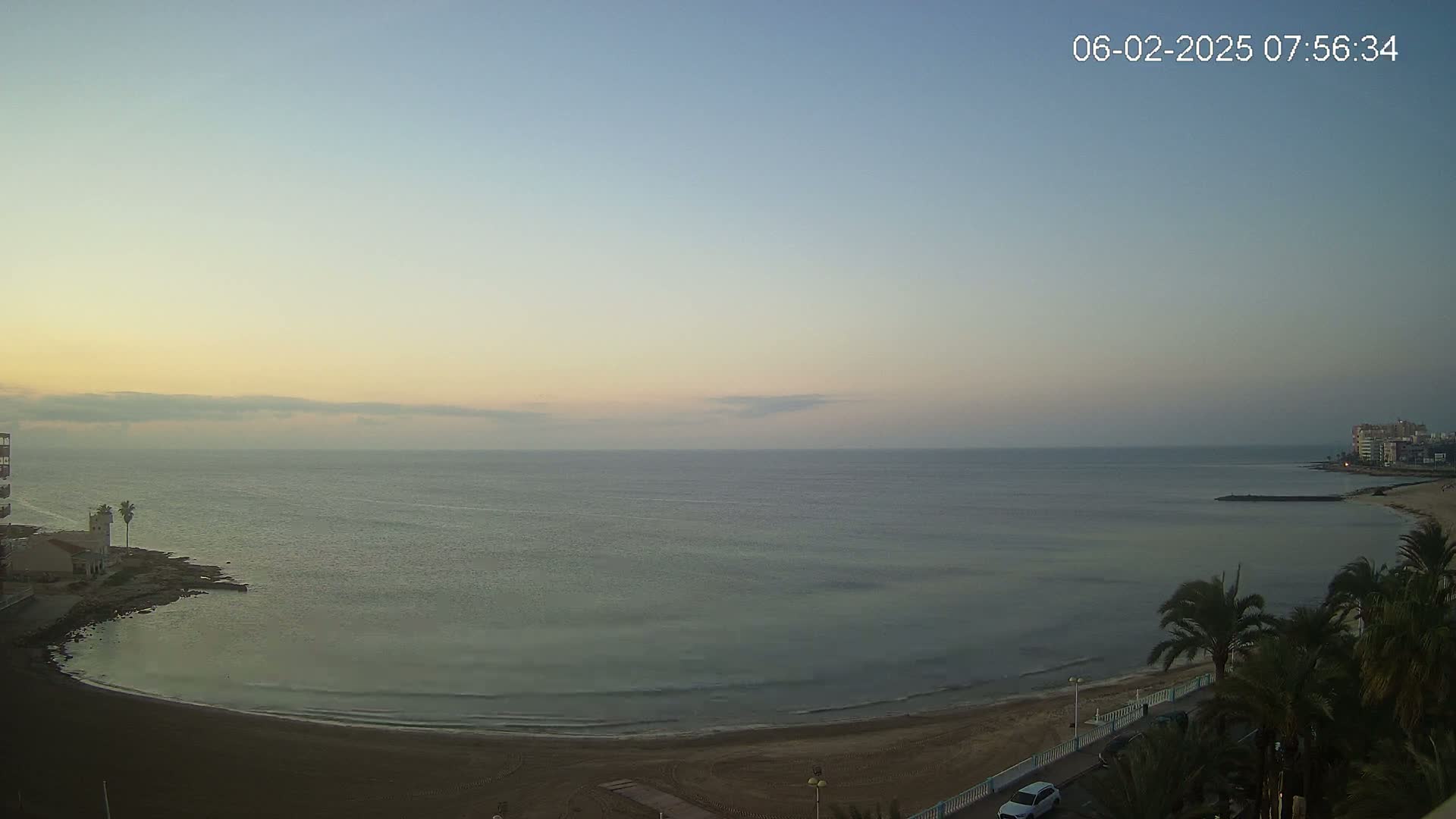 A calm, pale-blue sea laps a sandy beach at dawn under a mostly clear sky.