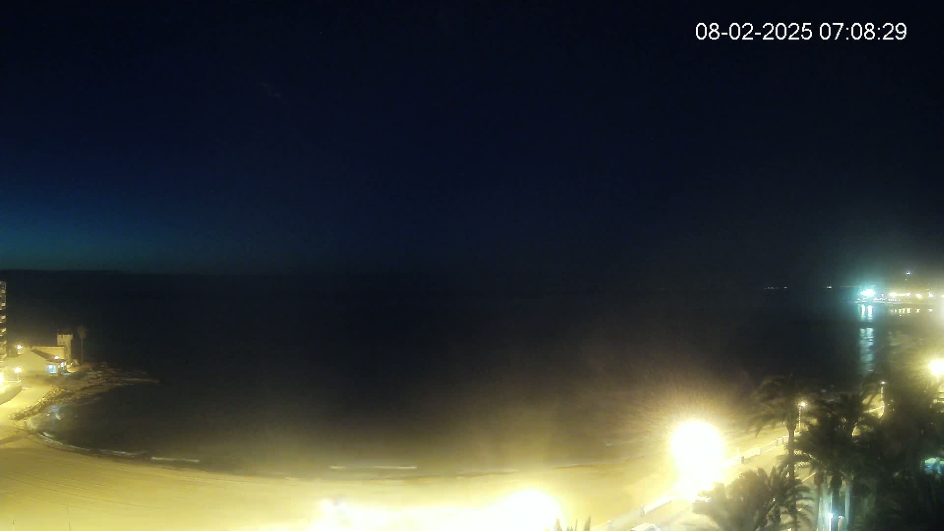 A nighttime view of a beach with calm waves, illuminated by streetlights, under a dark, clear sky.