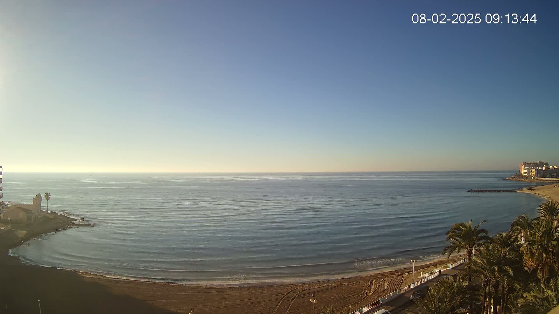 A sandy beach curves around a calm, blue ocean under a clear, bright sky.