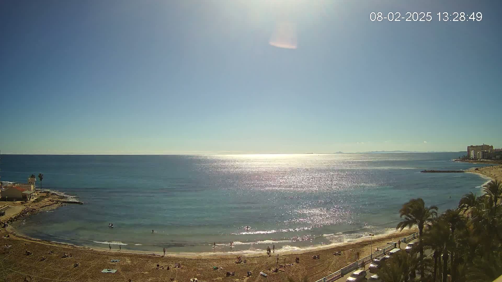 A sunny day at a beach with calm, sparkling water, a sandy shore dotted with people, and palm trees lining a nearby road.