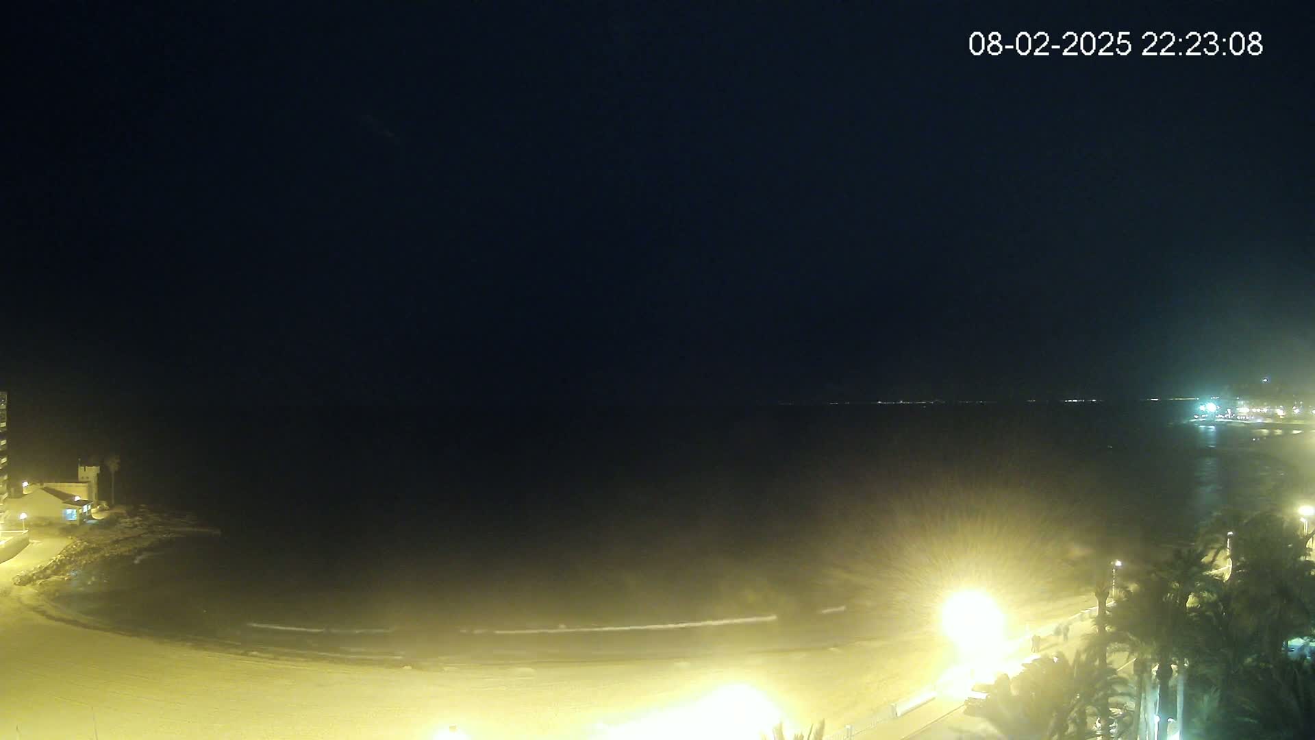 A nighttime view of a beach with gently lapping waves under a dark, clear sky, illuminated by artificial lights along the shore.