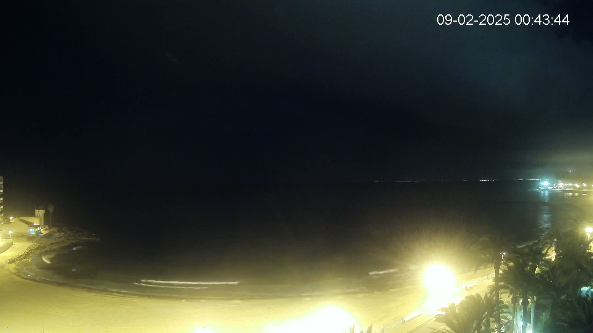 A nighttime view of a dimly lit beach with gentle waves lapping the shore, under a dark sky.