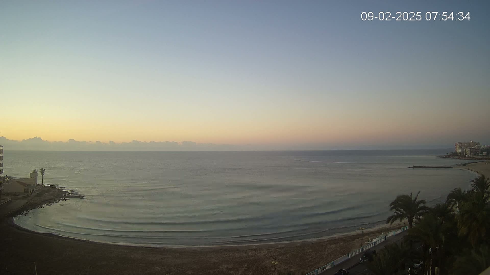 A calm, pale-pink sunrise illuminates a curved sandy beach and calm ocean water, with palm trees and buildings visible in the foreground and distance.