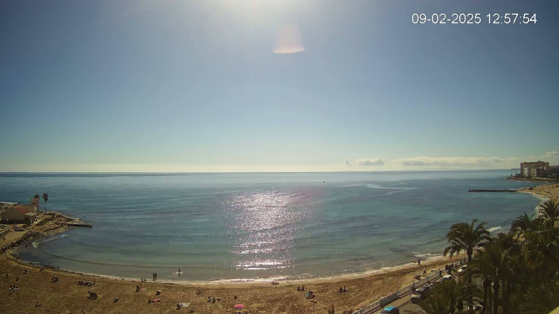 A sunny day at a sandy beach with calm, sparkling blue water, palm trees, and a few people scattered along the shoreline and in the water.