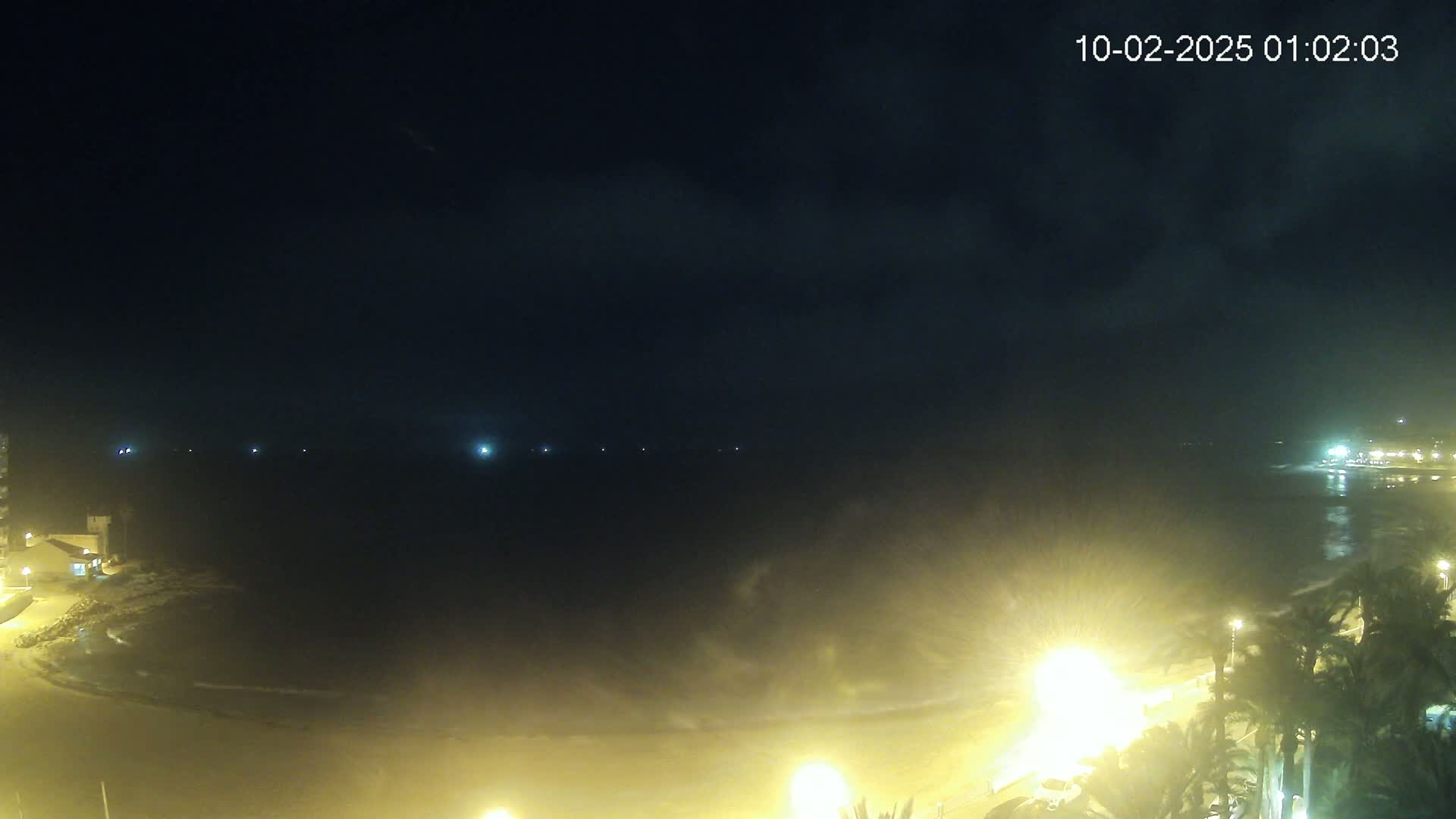 A nighttime view of a dark ocean shore illuminated by streetlights, under a mostly cloudy sky.