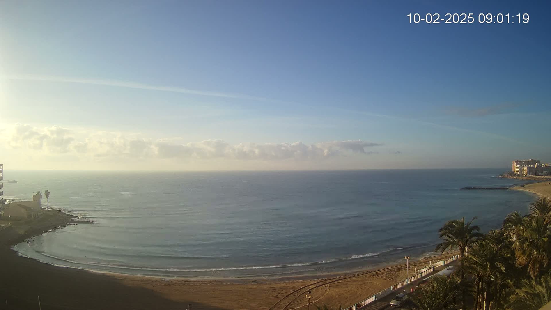 A calm, clear morning with a few clouds shows a sandy beach curving along a calm ocean, with buildings visible in the distance.