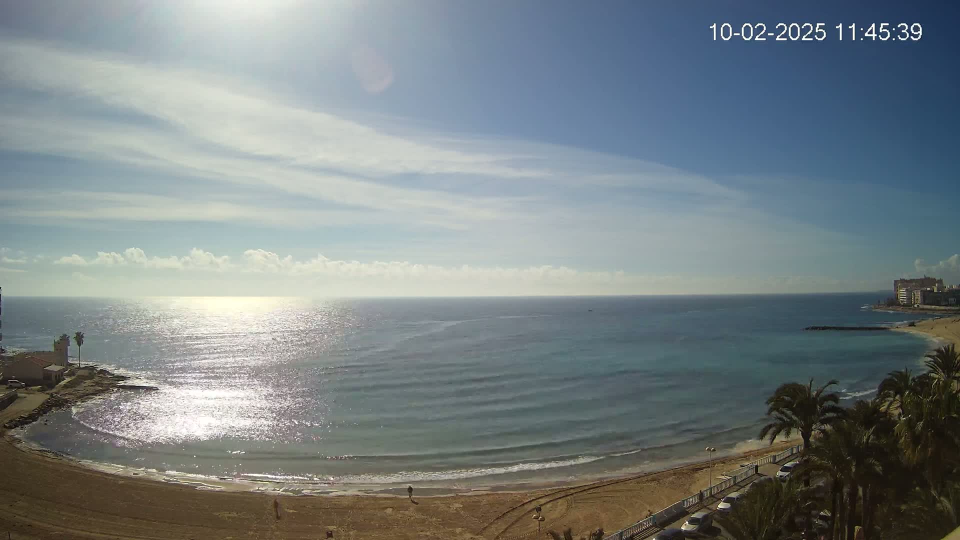 A sandy beach curves gently around a calm, sunlit sea under a mostly clear blue sky with scattered clouds.
