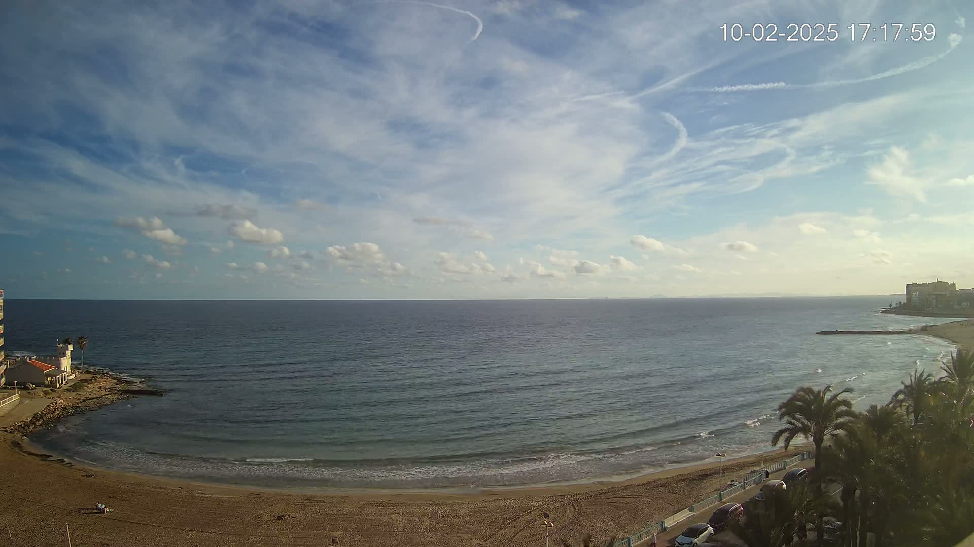 A mostly sunny day shows a calm ocean meeting a sandy beach, with palm trees and buildings visible on the shore.