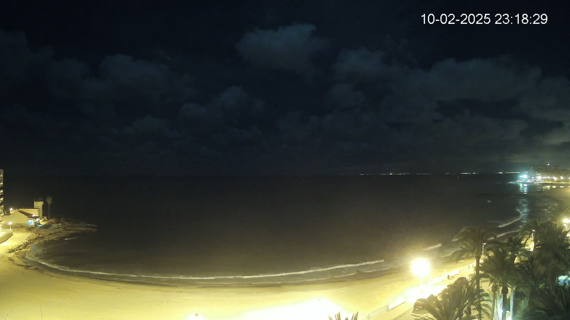 A nighttime beach scene shows gently lapping waves on a sandy shore under a partly cloudy sky, illuminated by streetlights and buildings.
