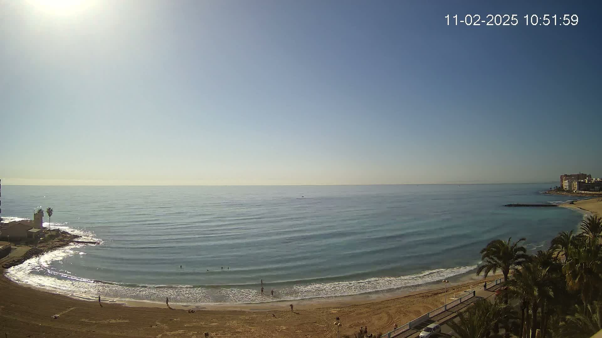 A sandy beach curves around a calm, blue ocean under a clear, sunny sky, with a few people visible on the shore and in the water.