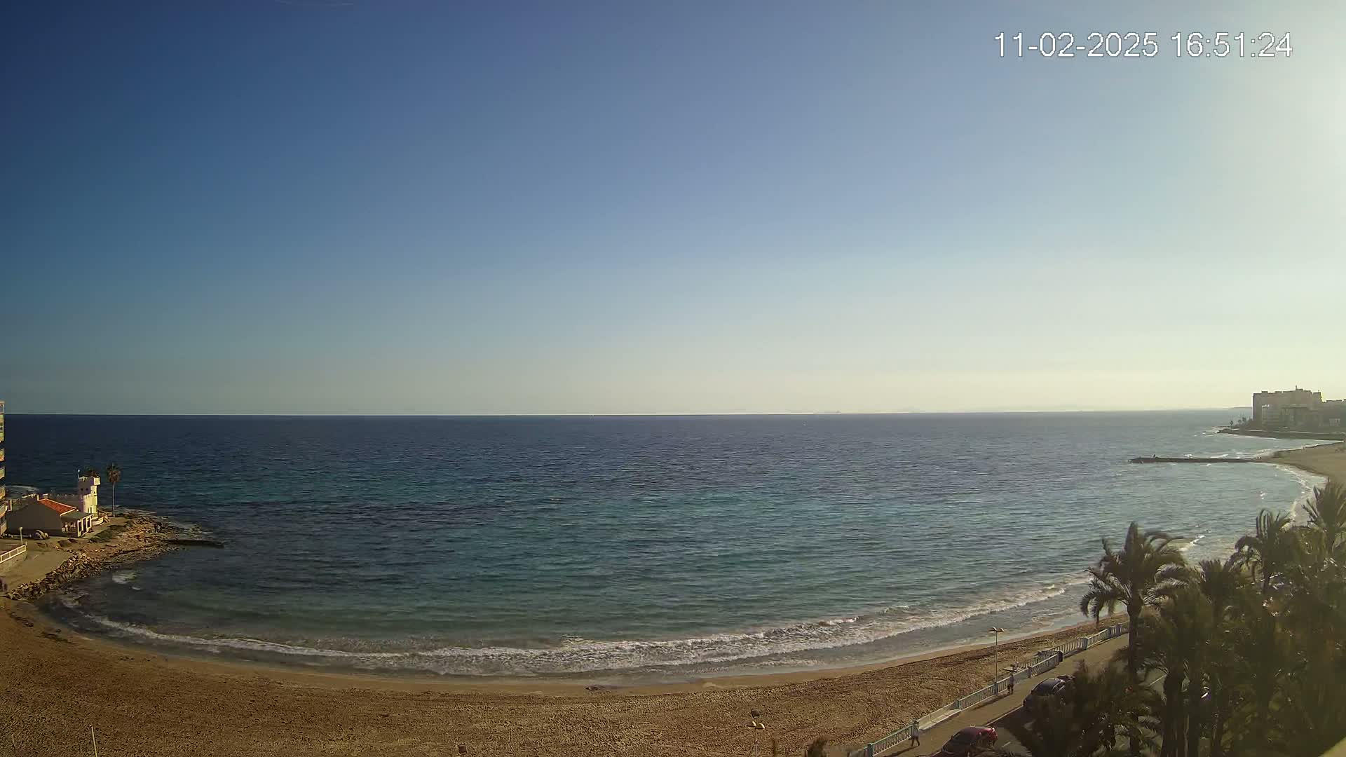 A sandy beach curves around a calm, clear blue sea under a clear blue sky.