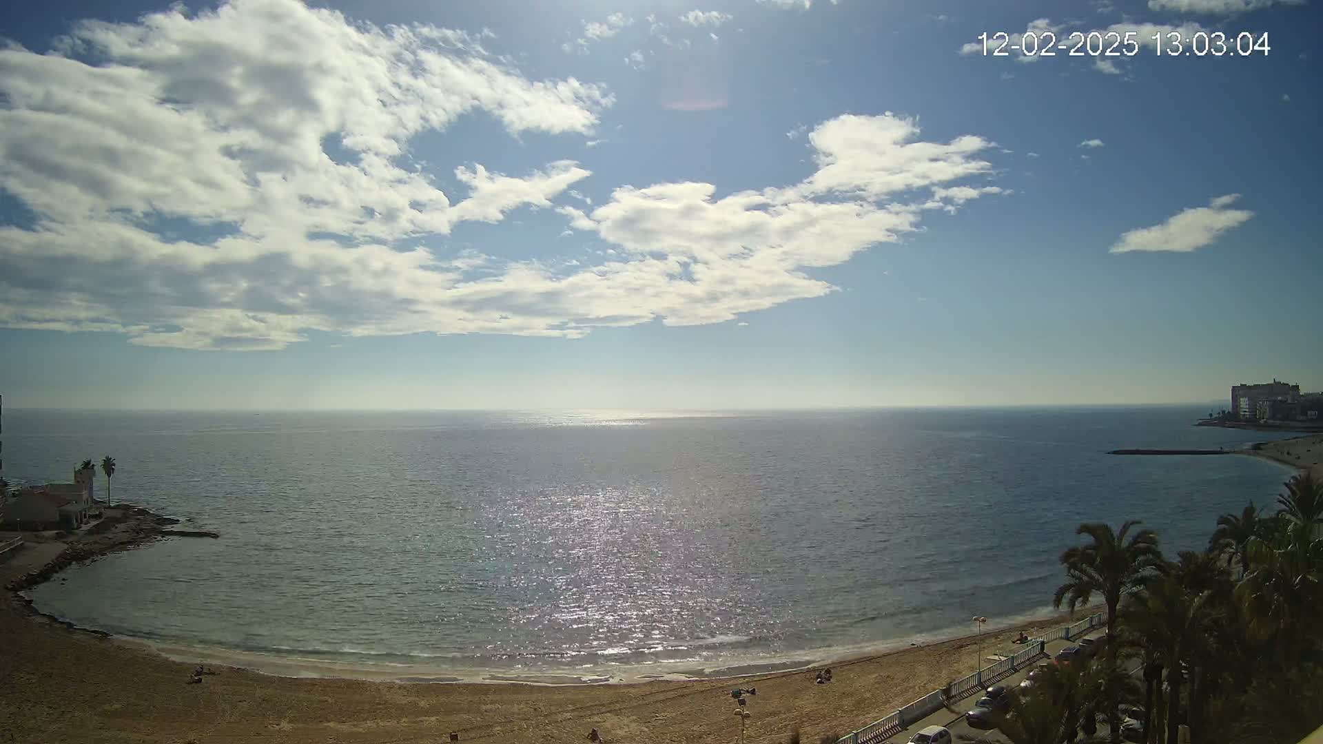 A mostly sunny day with scattered clouds shows a sandy beach curving into a calm sea, with palm trees and buildings visible in the distance.