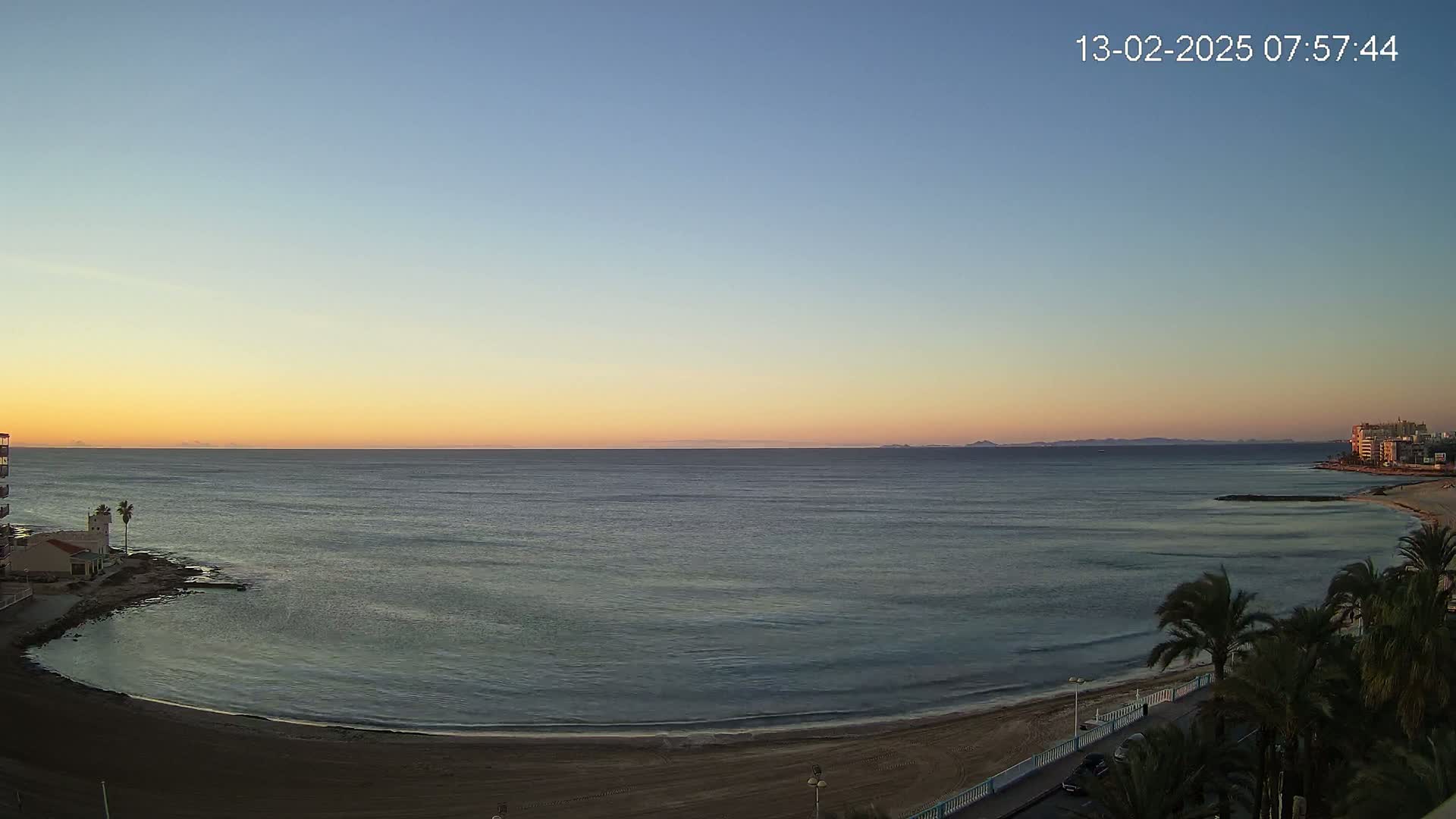 A calm ocean with a sandy beach and palm trees at sunset, under a clear sky.