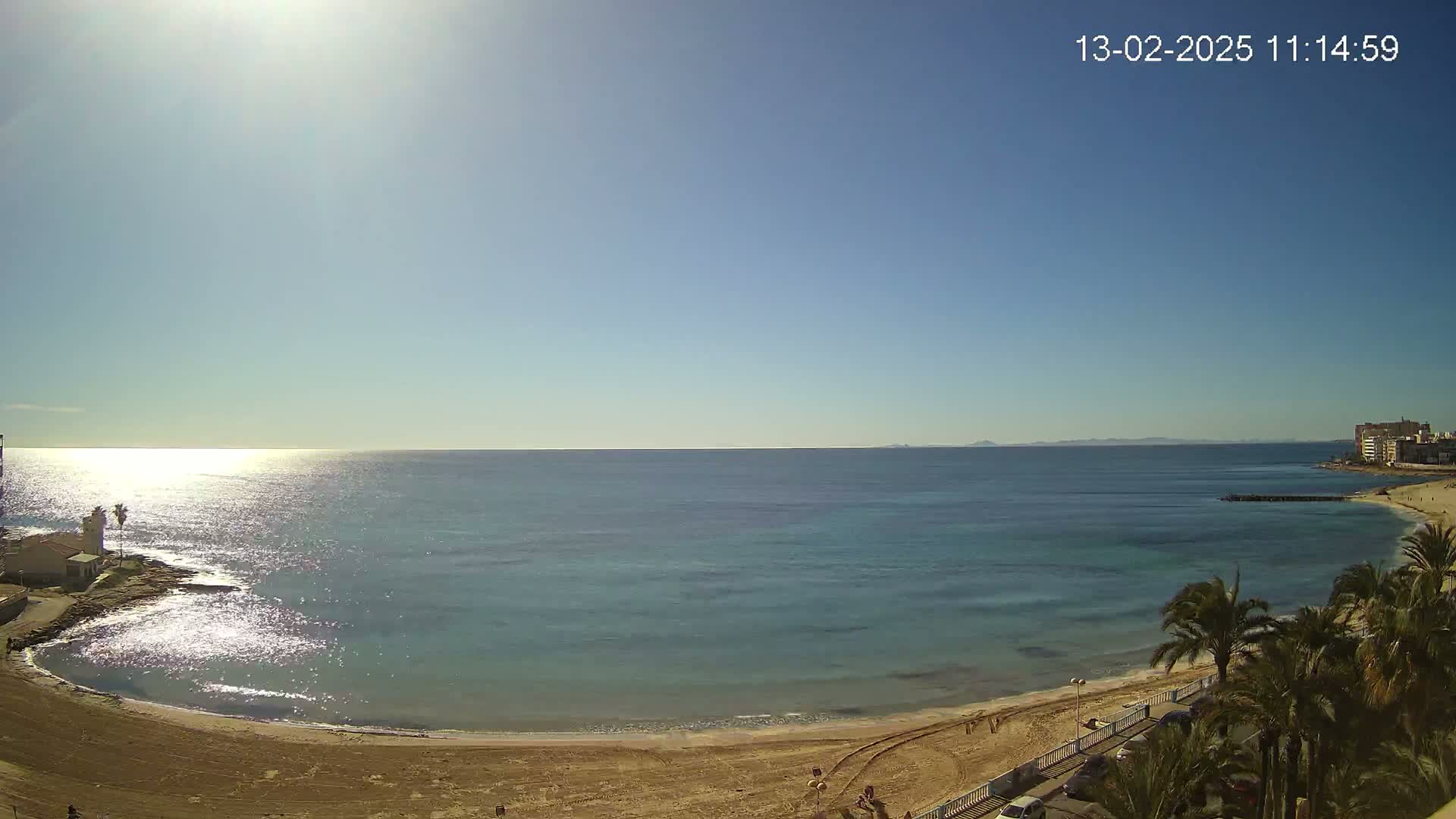 A sandy beach curves gently into a calm, sunlit turquoise sea under a clear blue sky.