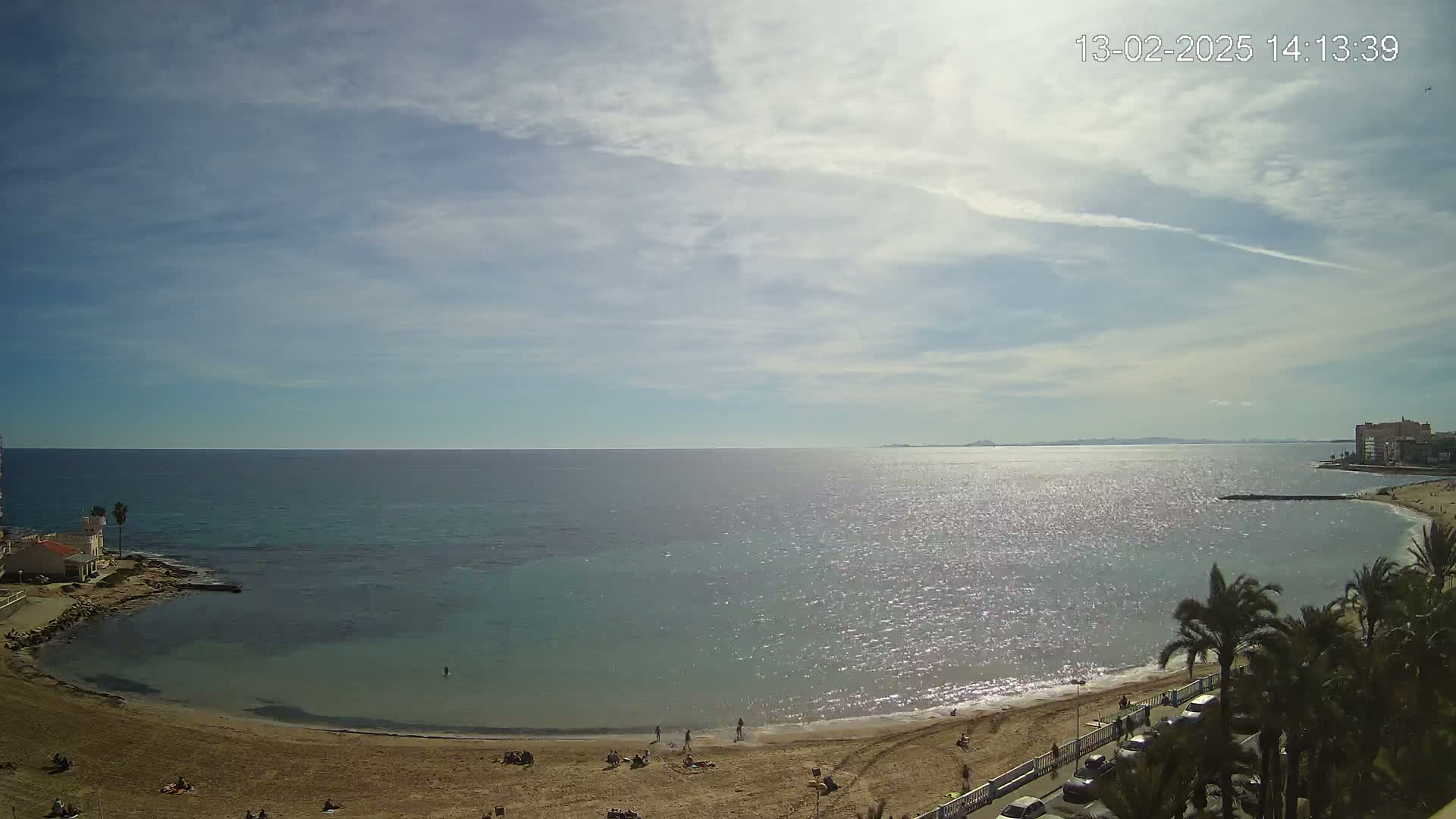 A sunny day at a beach with calm, sparkling water, a sandy shore with scattered people, palm trees, and buildings in the distance.