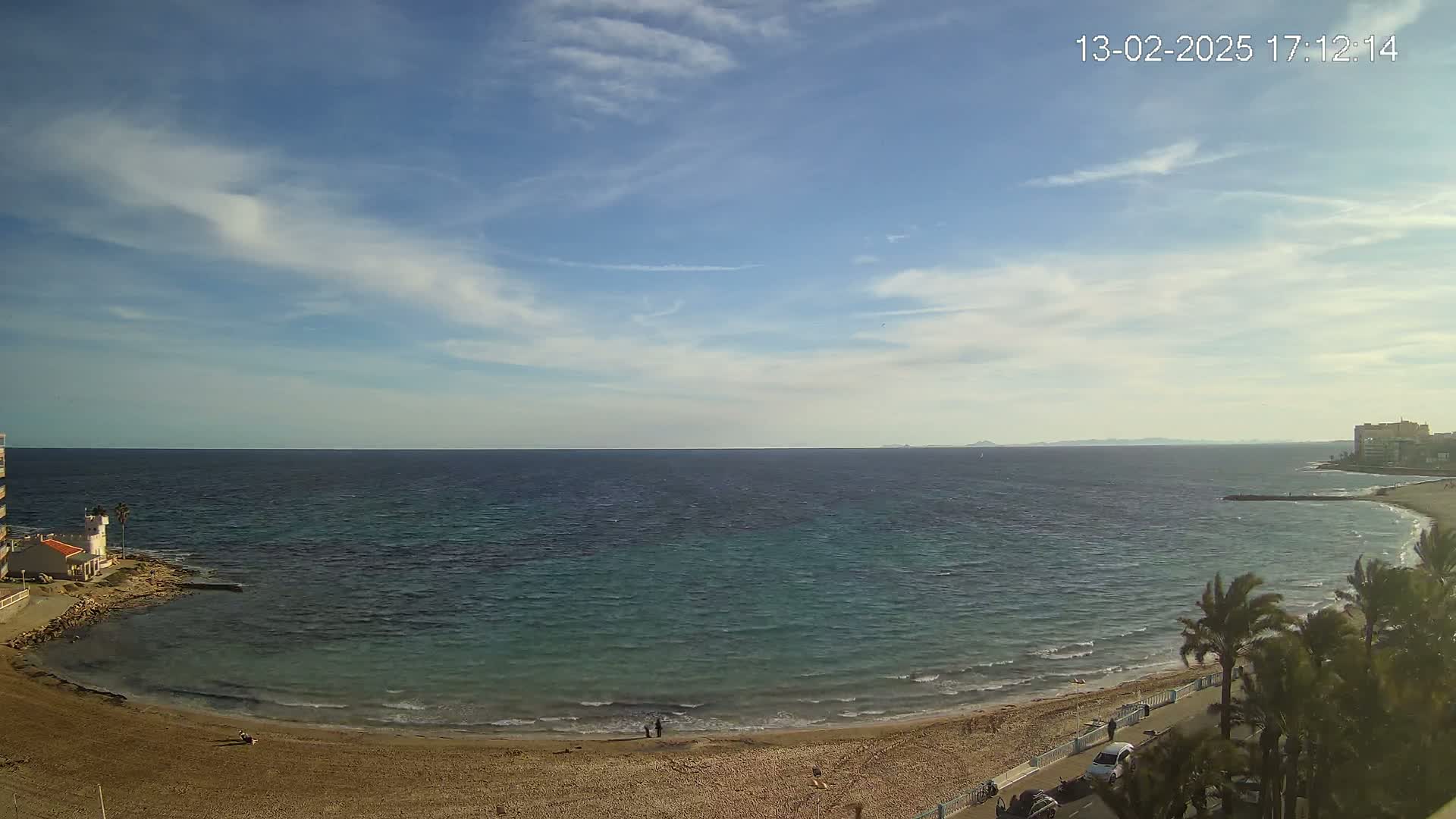 A sandy beach curves around a calm, teal-colored sea under a mostly clear, blue sky with a few scattered clouds.