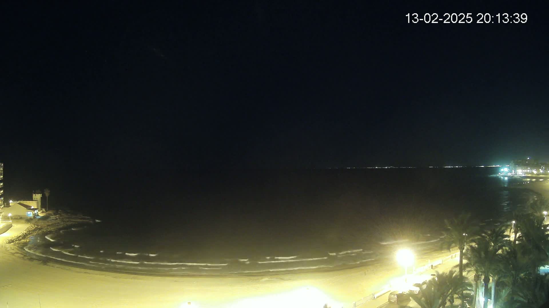A dark nighttime scene shows a gently waving sea lapping a sandy beach lined with palm trees and illuminated by streetlights.