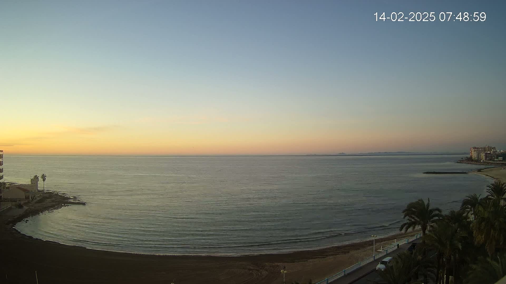 A calm, twilight ocean scene shows a sandy beach curving to the left, with palm trees and buildings in the foreground, and a distant coastline under a clear, pastel sky.