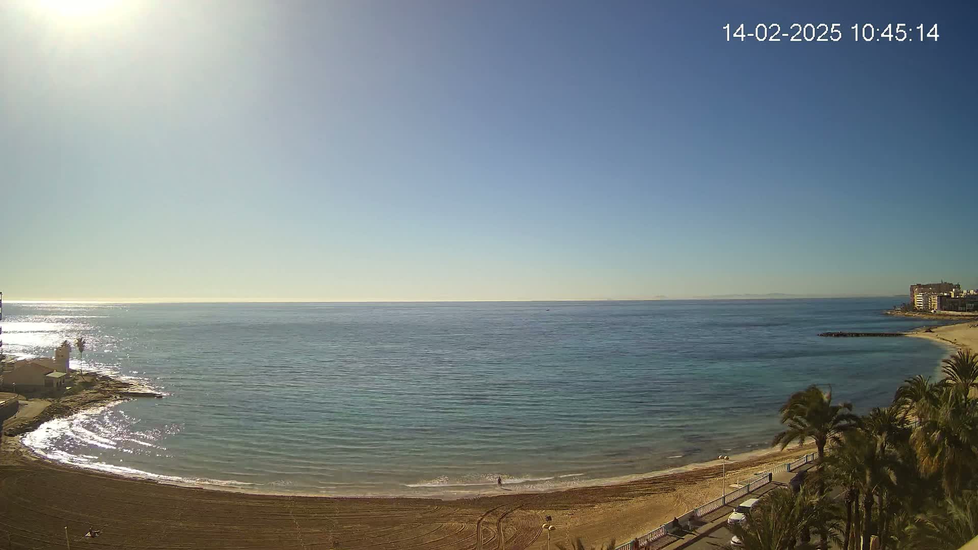 A sandy beach curves around a calm, clear ocean under a bright, sunny sky.