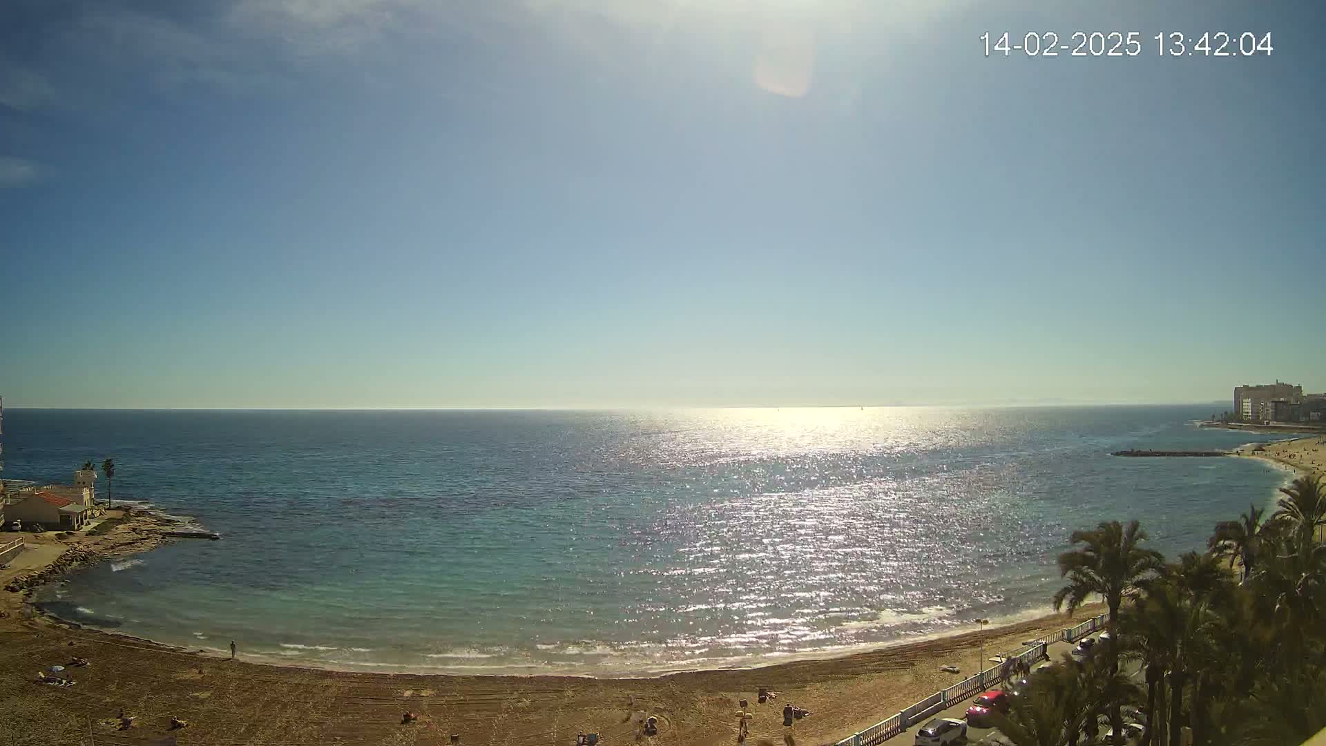 A sandy beach curves around a calm, sunlit ocean under a clear blue sky.