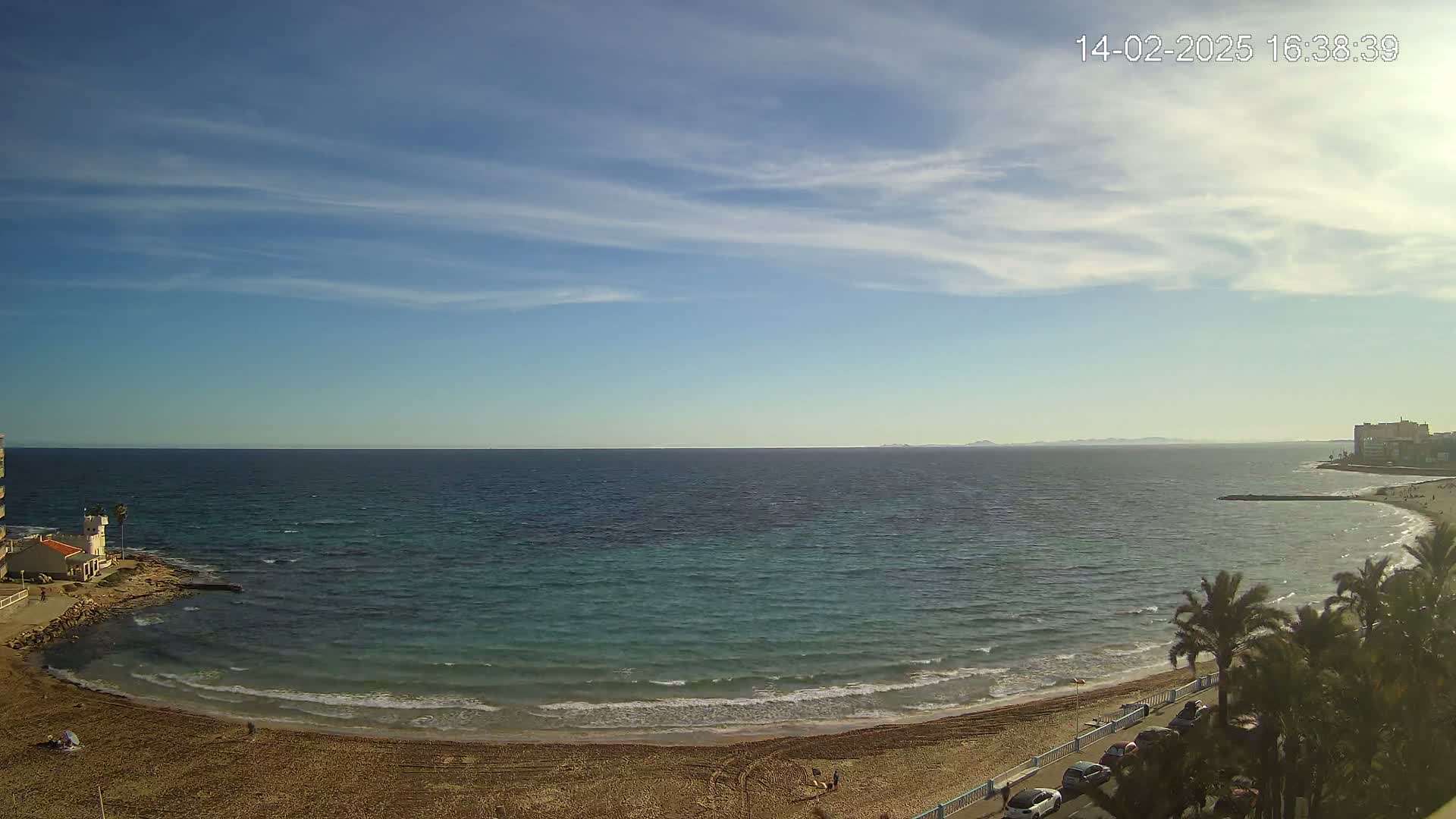 A sandy beach curves around a calm, teal-colored sea under a mostly clear, bright blue sky with thin, scattered clouds.