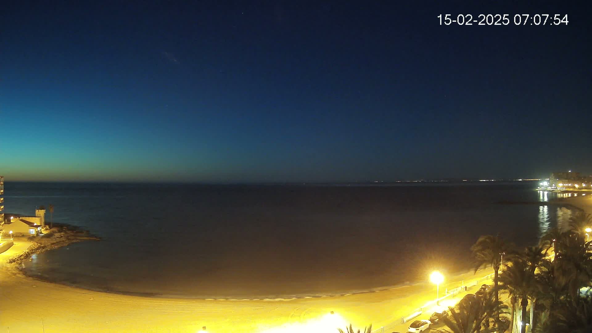 A dark-blue, pre-dawn sky overlooks a calm, dark-colored ocean, a sandy beach illuminated by artificial lights, and a palm tree-lined promenade.